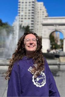 Picture of a smiling, bespectacled young woman with long, curly brown hair. She's wearing a purple hoodie and in front of the WSP fountain.