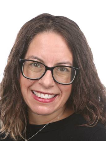 White-presenting woman with wavy, shoulder-length brown hair and chunky black glasses smiling at camera from a 3/4 profile