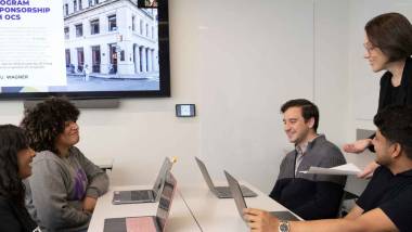 students sitting at table with adult standing handing out paper