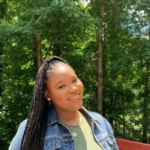 Niya is standing outside. There are trees with bright green leaves behind her. She is wearing a denim jacket with an olive green shirt underneath. 