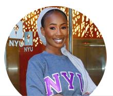 Fatima is standing in front glass doors with the NYU logo in white. Behind her is a red wall with lights. She is wearing a grey sweatshirt and NYU in purple. She is smiling. 